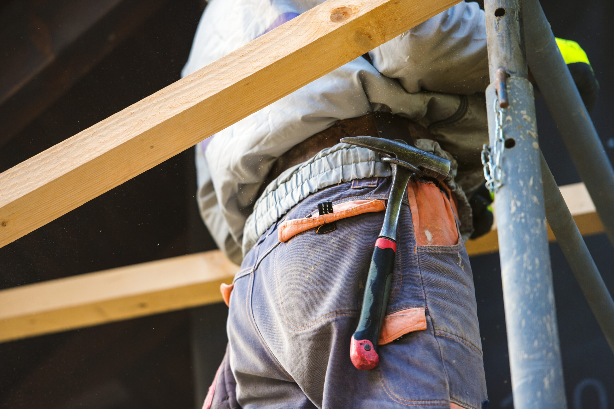 A construction worker stands on scaffolding, wearing work pants with a hammer in the back pocket. Wooden beams and metal poles are visible, indicating building or repair work in progress.